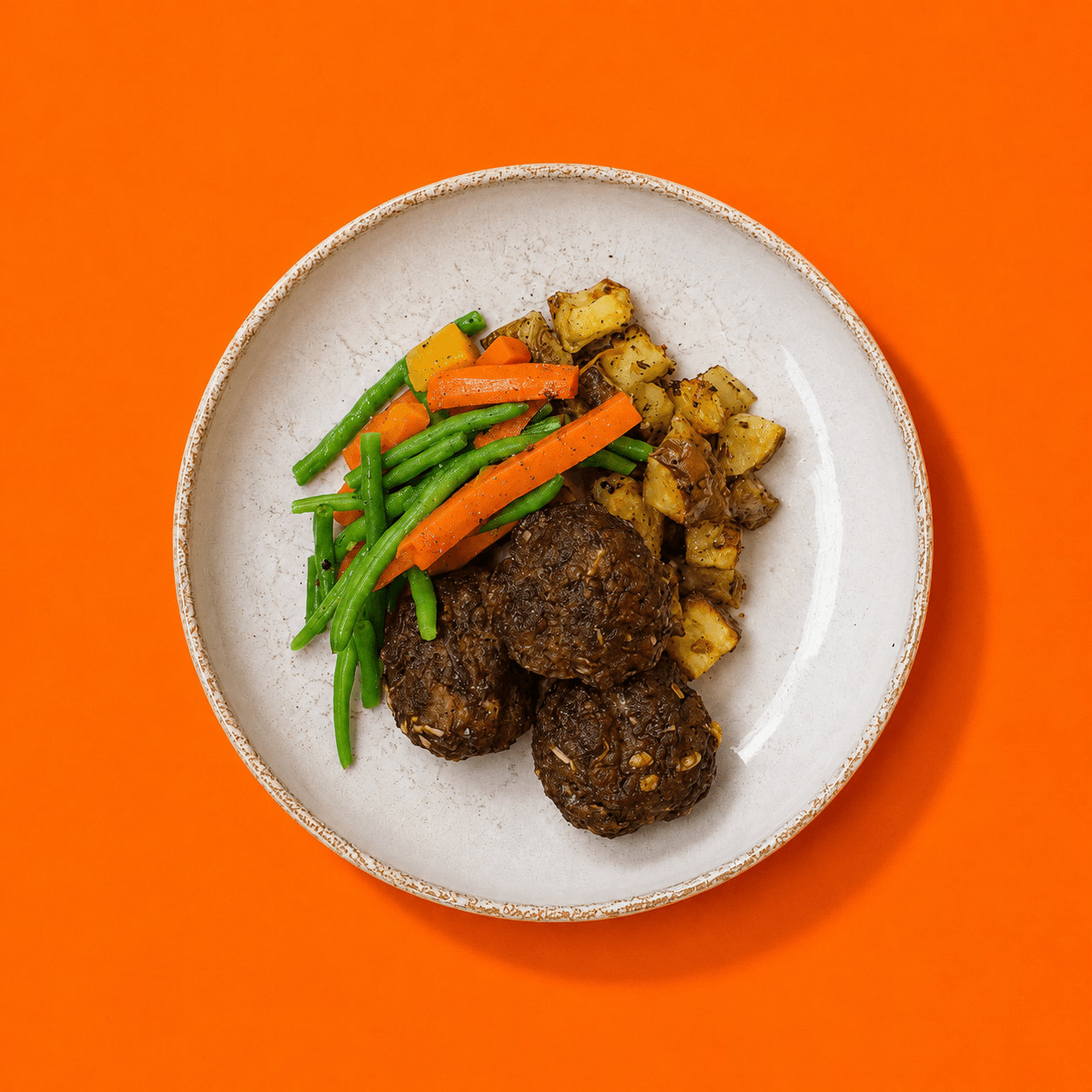Assiette de boulettes de viande, pommes de terre rôties, haricots verts et carottes, photographiée du dessus sur un fond orange vif. Bigmig Quebec