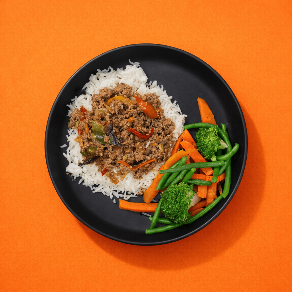 Une assiette noire mate contient du riz blanc, un sauté aux poivrons et oignons, accompagné de légumes verts (brocoli, haricots verts) et de carottes. Bigmig prêt-à-manger sans gaspillage